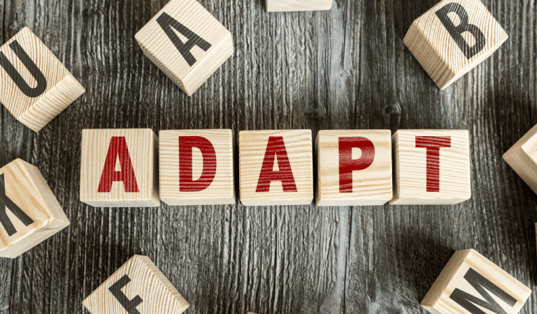 The word adapt in red text printed on 5 wooden blocks on a wooden table with other blocks around it.